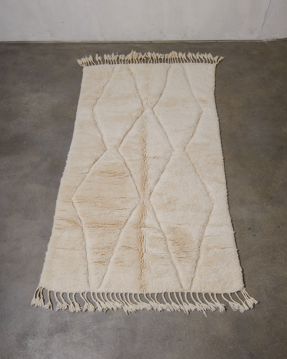 Beige woven rug with diamond pattern and fringe on a concrete floor.
